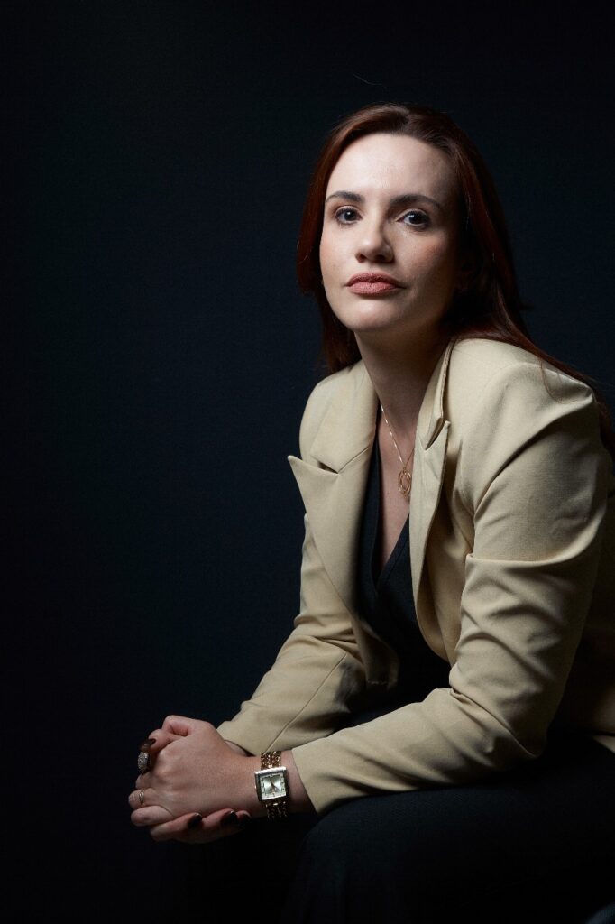 Doutora Ynara Nieto Mulher de cabelos castanhos avermelhados, usando blazer bege e blusa preta, sentada com as mãos cruzadas sobre o colo, em um fundo escuro, olhando diretamente para a câmera com expressão séria